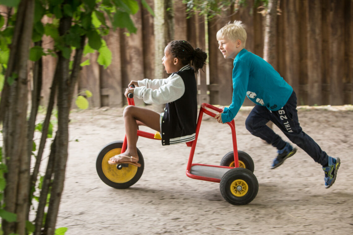 dreieins Innovative Pädagogik – dreieins Kindertagesstätte Weissensee Kinder spielen im Garten: Ein Kind sitzt auf einem Dreirad und ein anderes Kind schiebt das Dreirad an.
