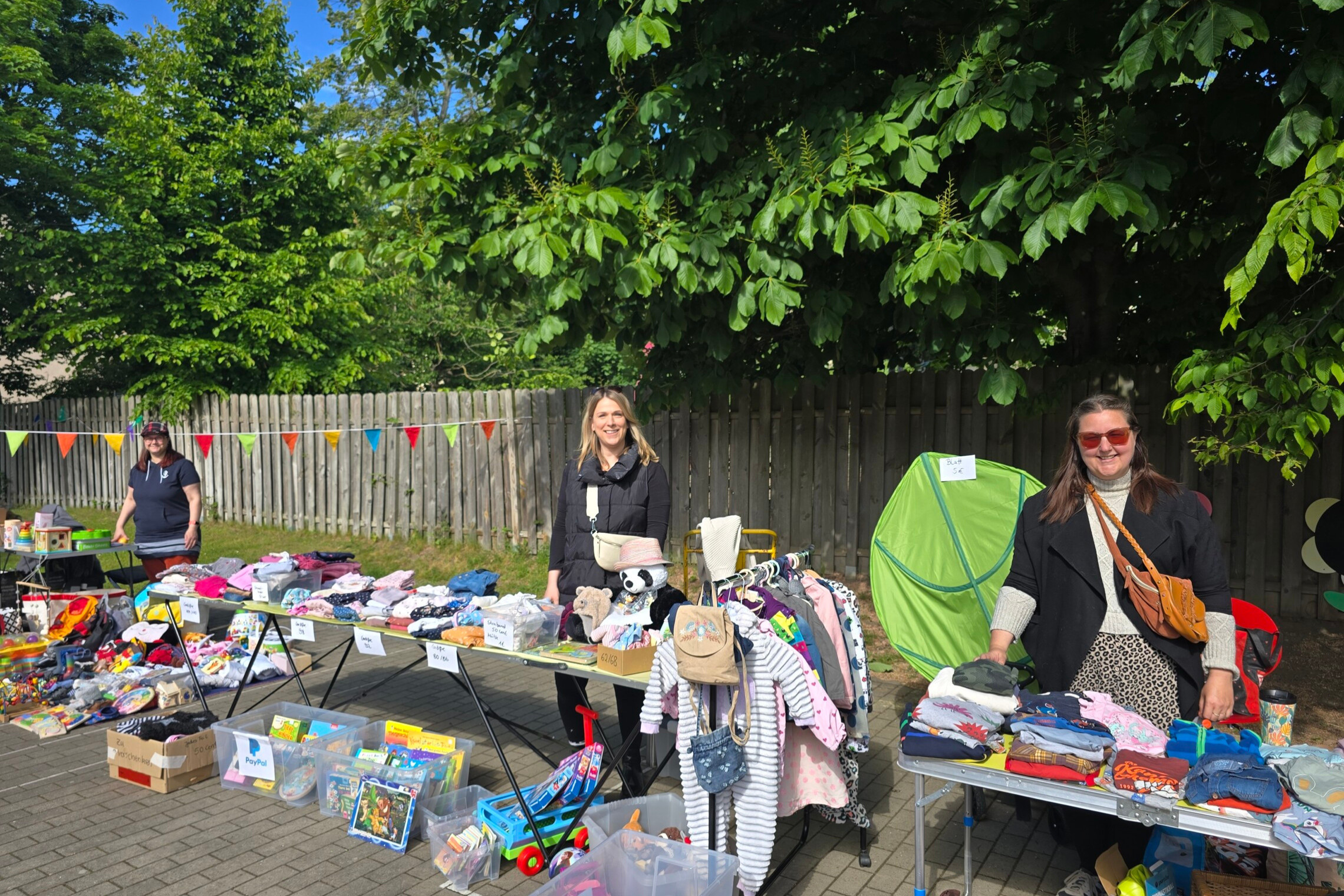 dreieins Kindertagesstätte Prenzlauer Berg - Flohmarkt in der Kita: Auf dem Bild zu sehen, sind Menschen, die hinter Tischen stehen, auf denen Kleidung, Bücher und Spielzeug liegen.