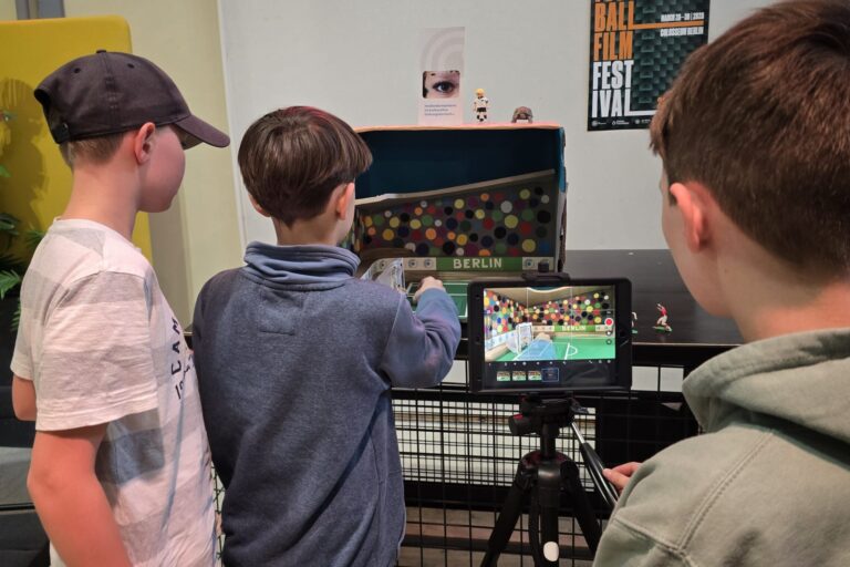dreieins Schüler filmen eine Fußballszene an einem Setmodell